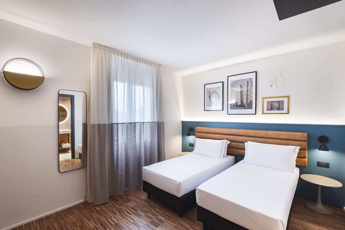 A hotel room with two beds, a round mirror, a bedside table, and a wall-mounted lamp.