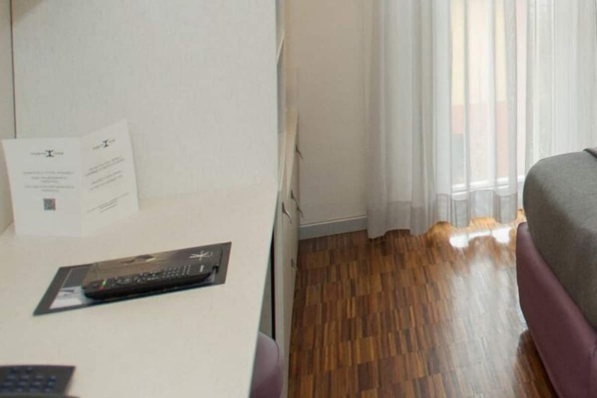 A hotel room with a bed, a bedside table with a remote control, a window with curtains, and a patterned wall.