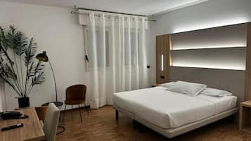 A modern hotel room with a wooden floor, a bed with white linens, a desk with a chair, a potted plant, and a large window with white curtains.