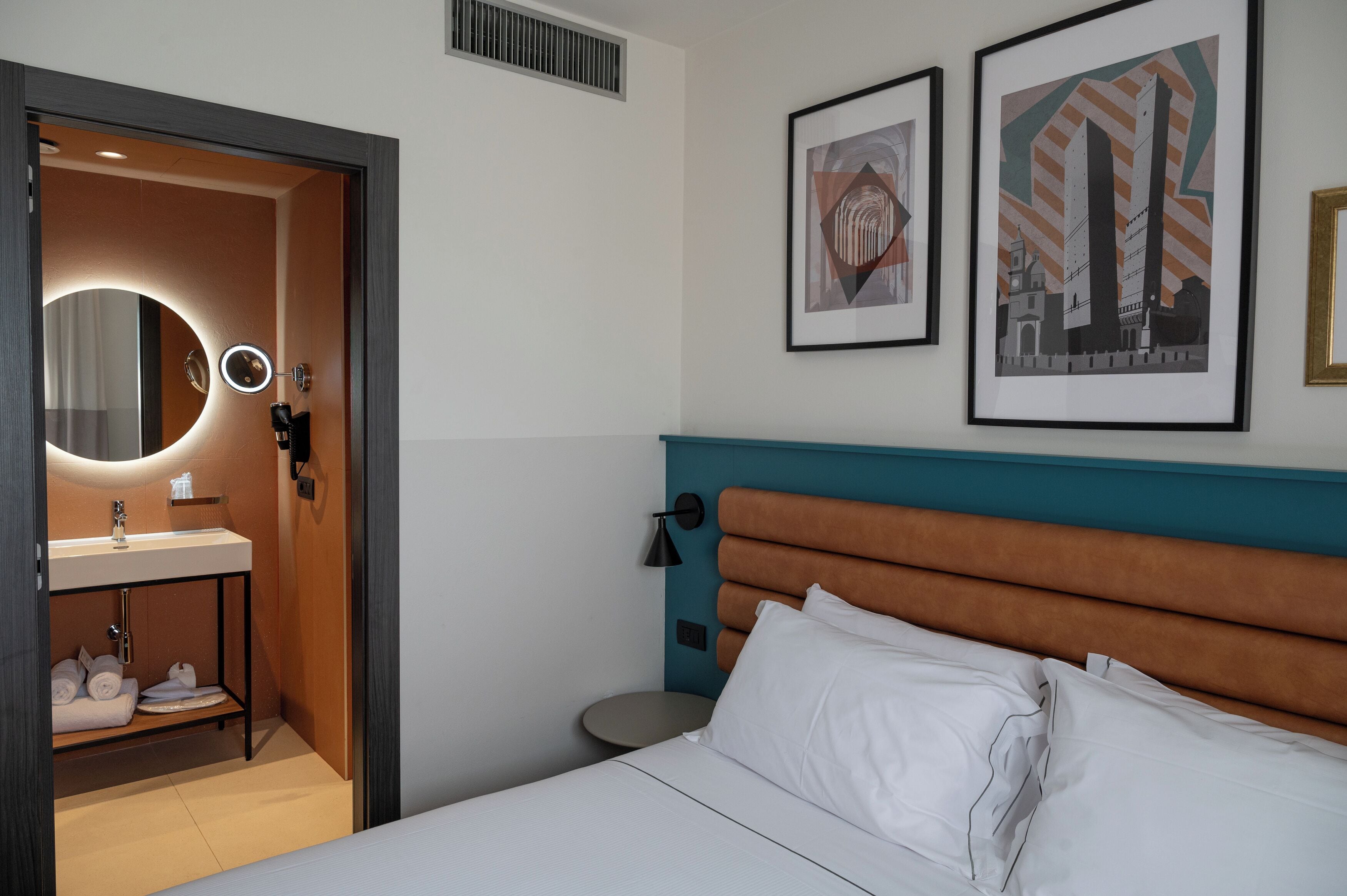 Habitación de hotel con una cama, un espejo redondo, un lavamanos y dos cuadros en la pared.