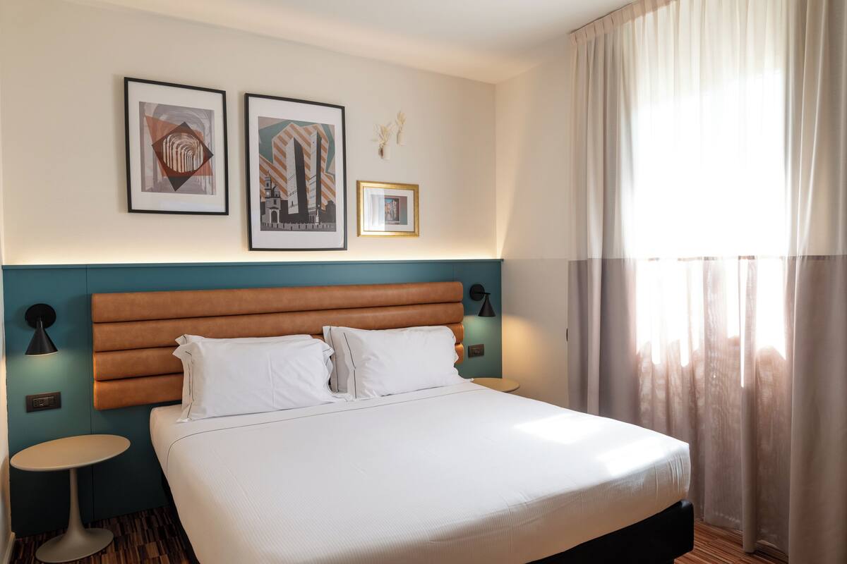 A hotel room with a large bed, two framed pictures on the wall, a nightstand, and a window with curtains.