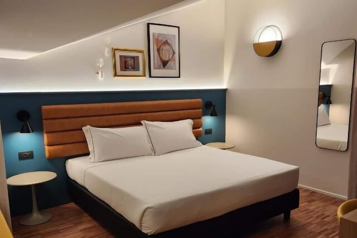 A modern hotel room with a large bed, a wooden headboard, a small round table, and a wall-mounted mirror.
