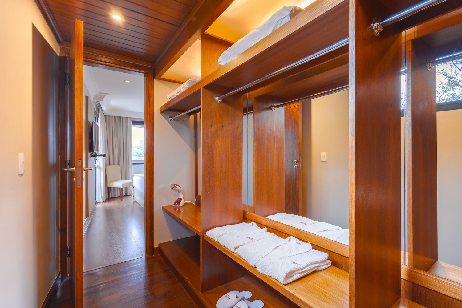 A hotel room with a wooden wardrobe, a mirror, a shelf with folded towels, and a small table with a bottle.