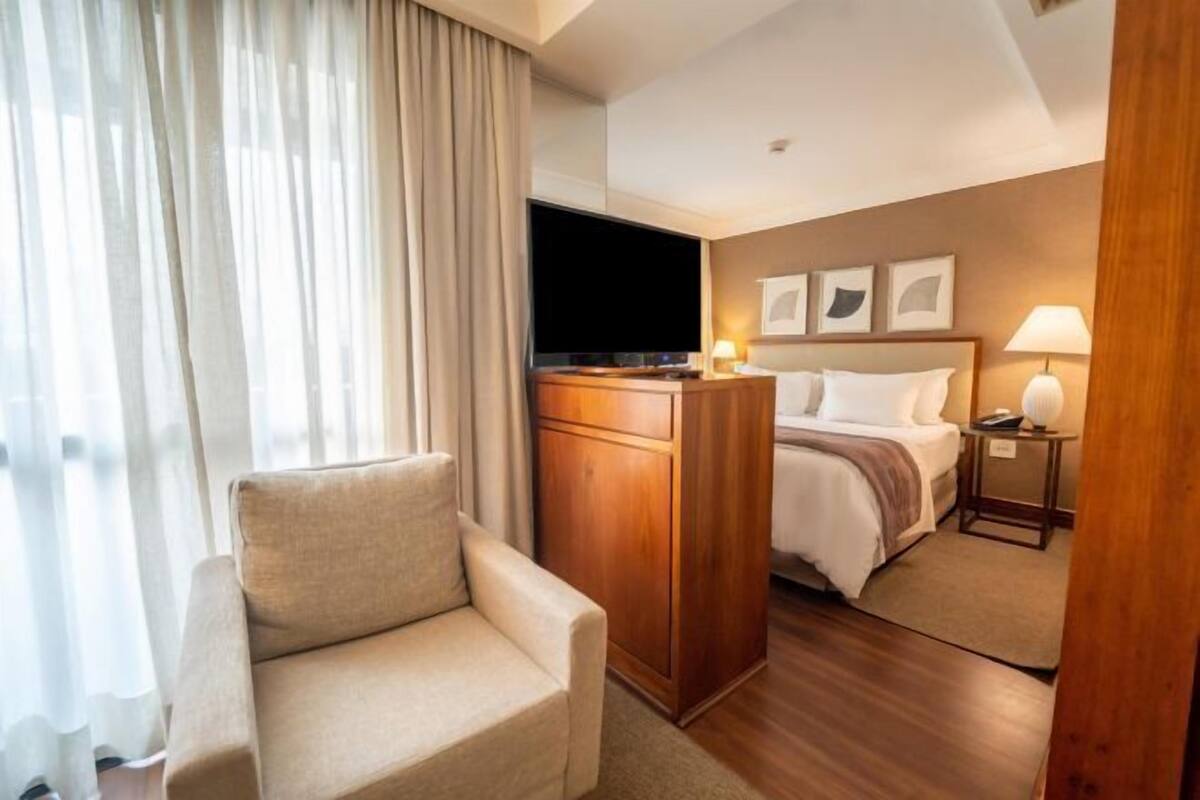 A hotel room with a bed, a desk, a chair, a TV, and a window with curtains.