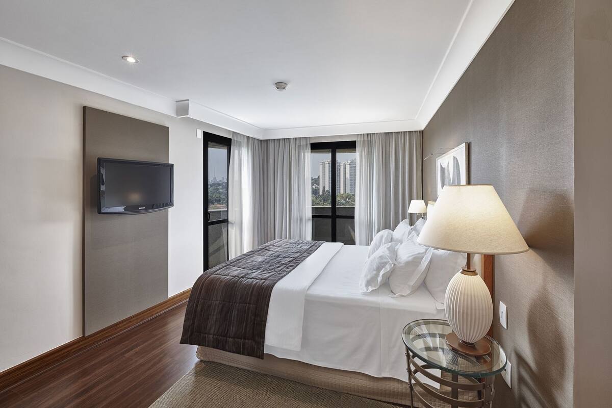 A hotel room with a bed, a TV, a lamp, and a view of the city.
