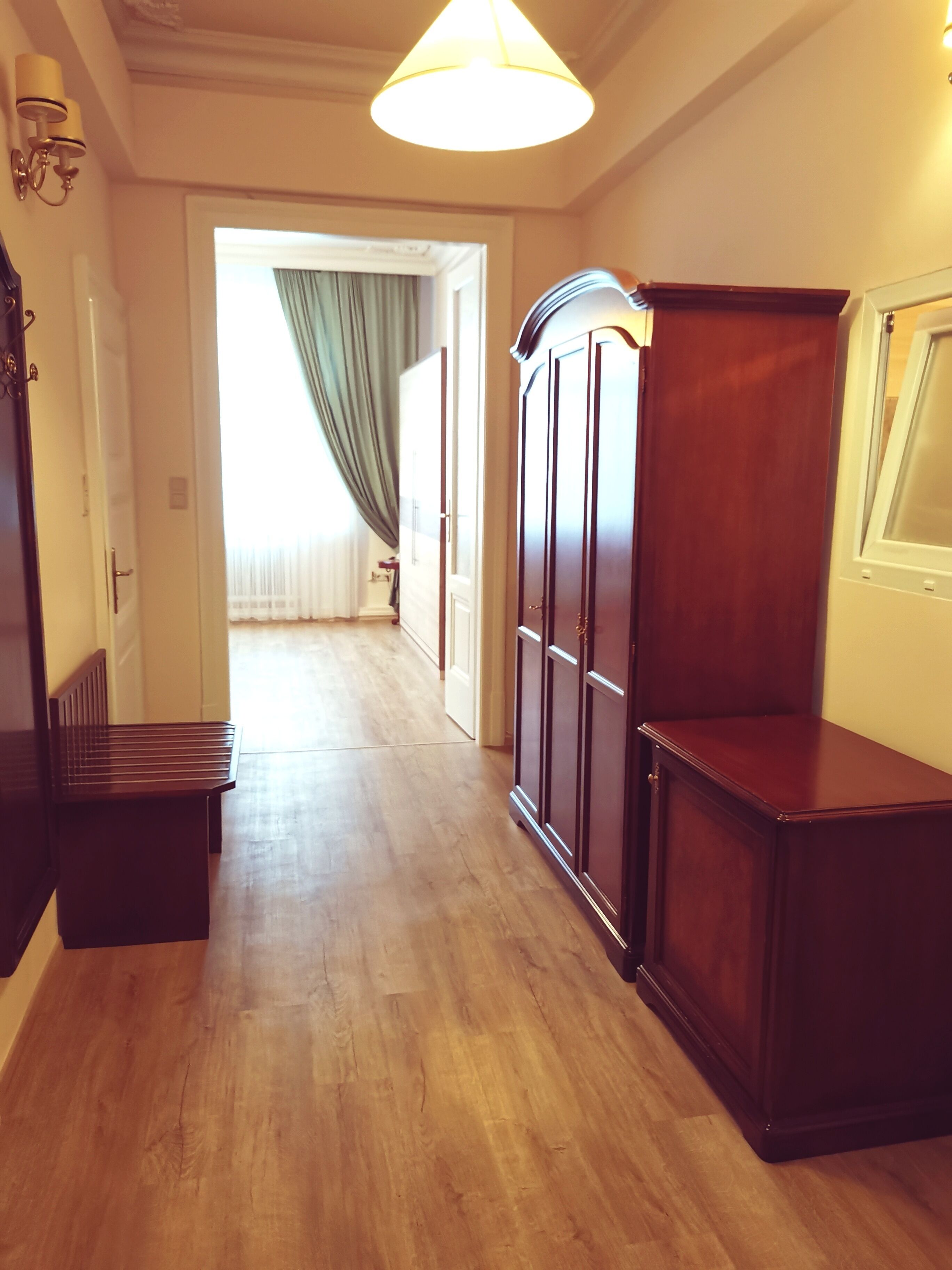 A hallway with wooden flooring, a bench, a wardrobe, and a door leading to another room with a window and curtains.