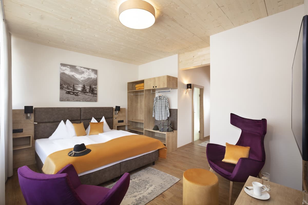 Ein modernes Hotelzimmer mit einem großen Bett, einem Holzkopfende und einem Gemälde an der Wand. Es gibt violette Sessel, einen kleinen Tisch mit einer Tasse und einen Fernseher an der Wand.