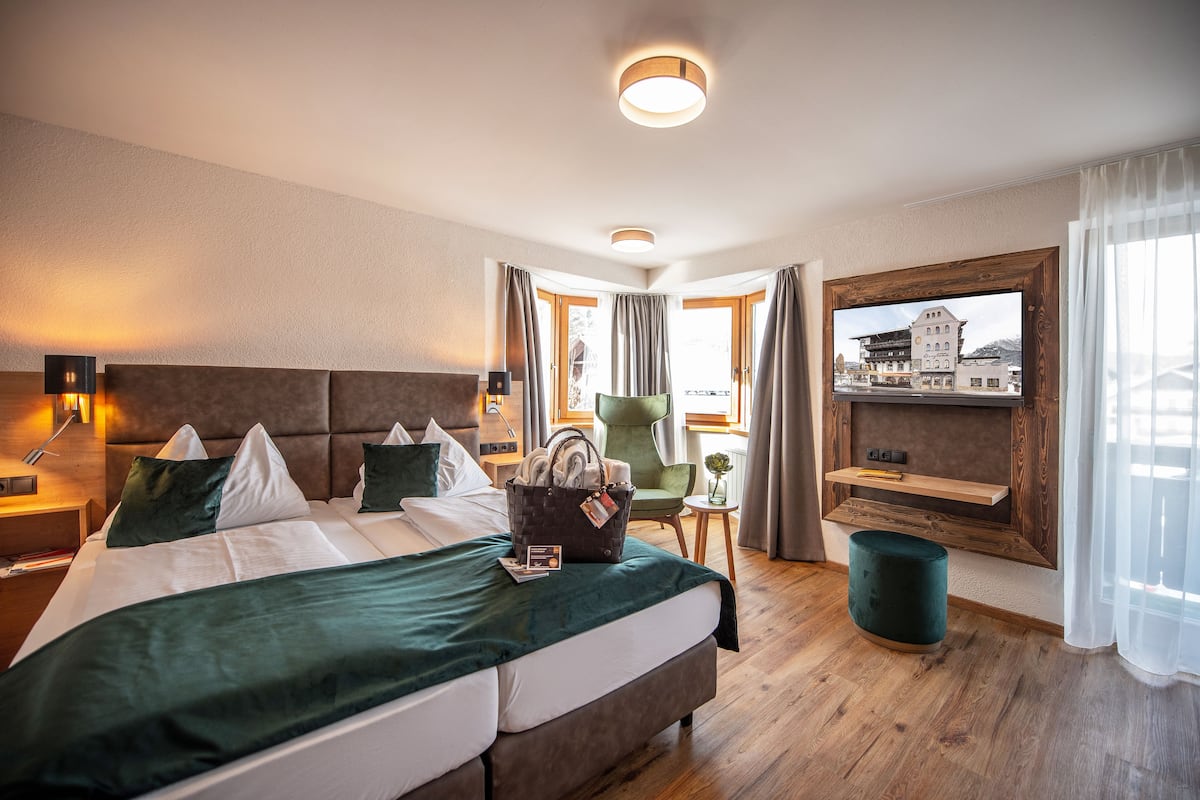 Ein Hotelzimmer mit einem großen Bett, einem Sessel, einem kleinen Tisch, einem Fernseher und einem Fenster mit Vorhängen.