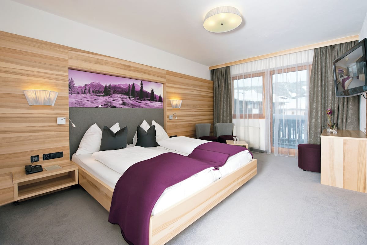 Ein modernes Hotelzimmer mit einem großen Bett, einem hölzernen Kopfteil, zwei Nachttischlampen, einem Flachbildfernseher, einem kleinen Schreibtisch und einem großen Fenster mit Vorhängen.
