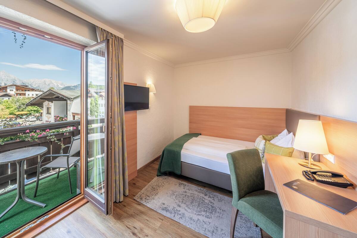 Ein Hotelzimmer mit Bett, Schreibtisch mit Lampe, Stuhl, Fernseher und Balkon mit Tisch und Stühlen.