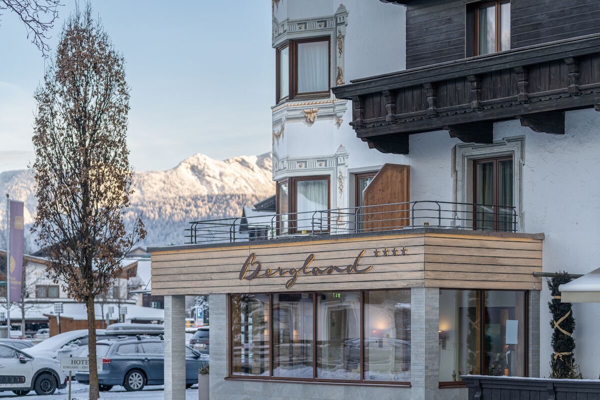 Un hotel llamado 'Bergland' con balcón, ventanas amplias y vistas a montañas nevadas.