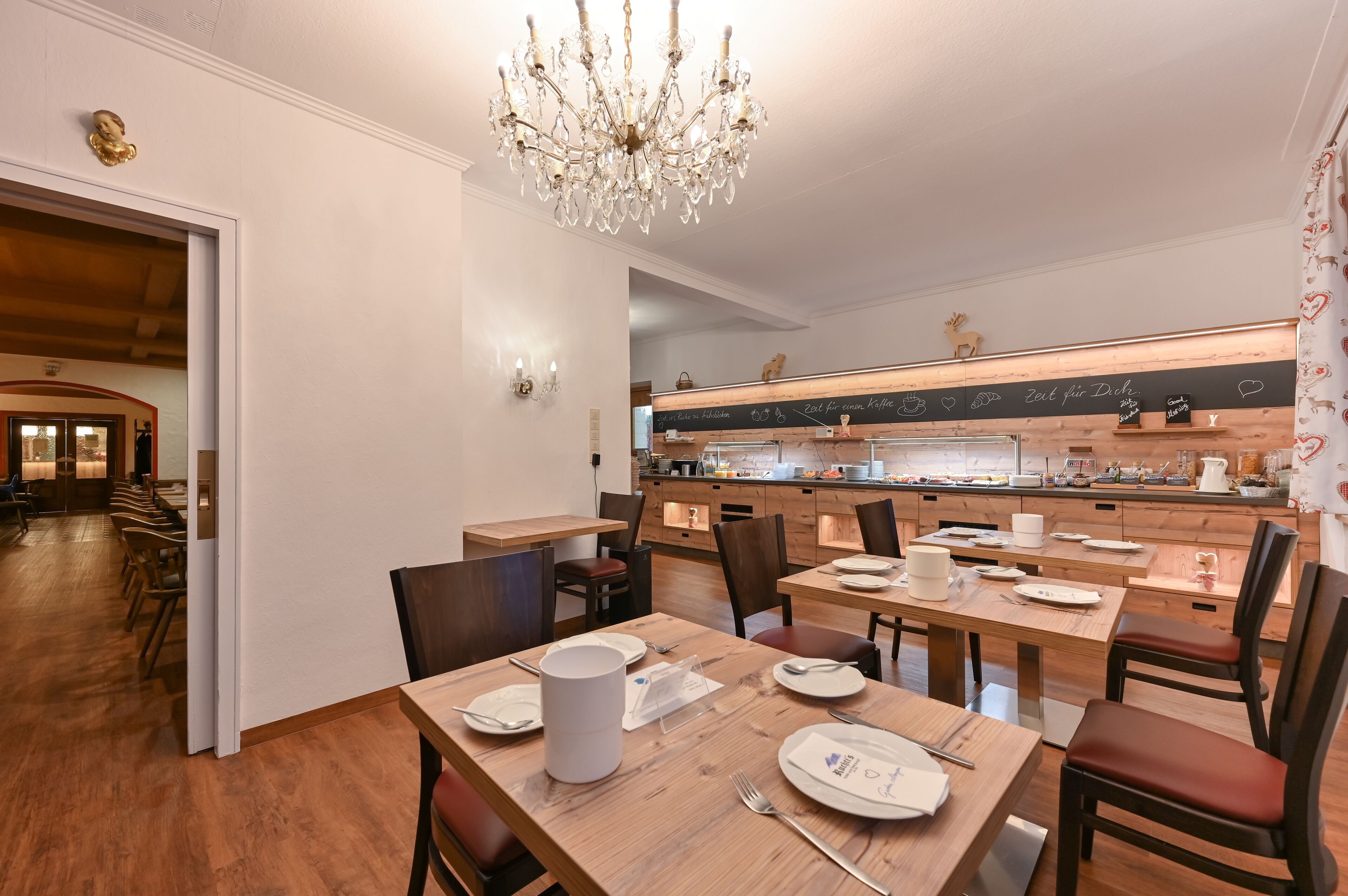 Un comedor con mesas y sillas de madera, una araña y una cocina al fondo.