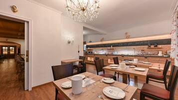 Un comedor con mesas y sillas de madera, una araña y una cocina al fondo.