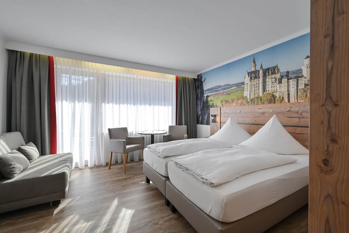 A hotel room with a bed, a sofa, a desk, and a chair. There is a large window with curtains and a painting of a castle on the wall.