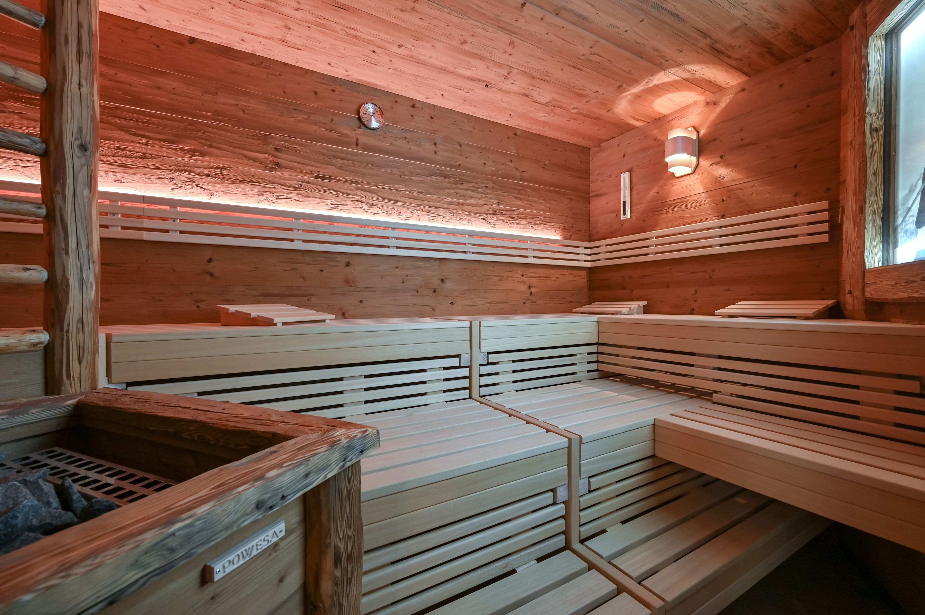 A sauna with wooden benches and a stone basin, featuring a sloped ceiling and a wall-mounted light fixture.