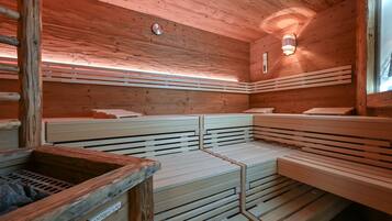 A sauna with wooden benches and a stone basin, featuring a sloped ceiling and a wall-mounted light fixture.