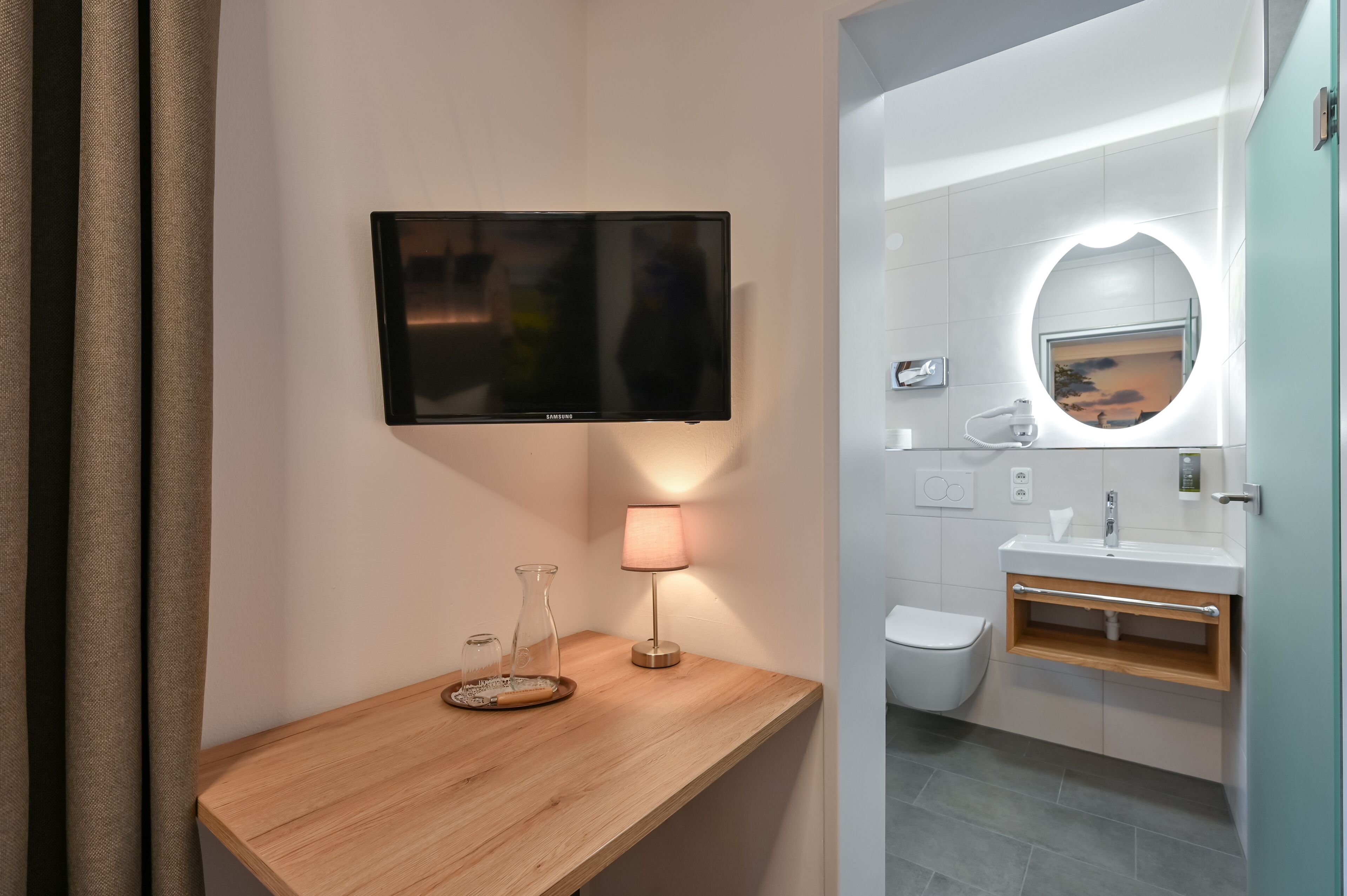 A modern bathroom with a wooden vanity, a flat-screen TV, a lamp, a mirror with a circular light, and a toilet.