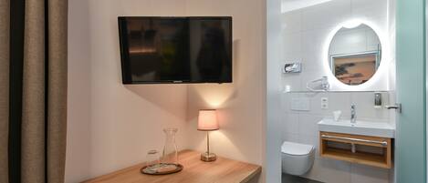 A modern bathroom with a wooden vanity, a flat-screen TV, a lamp, a mirror with a circular light, and a toilet.