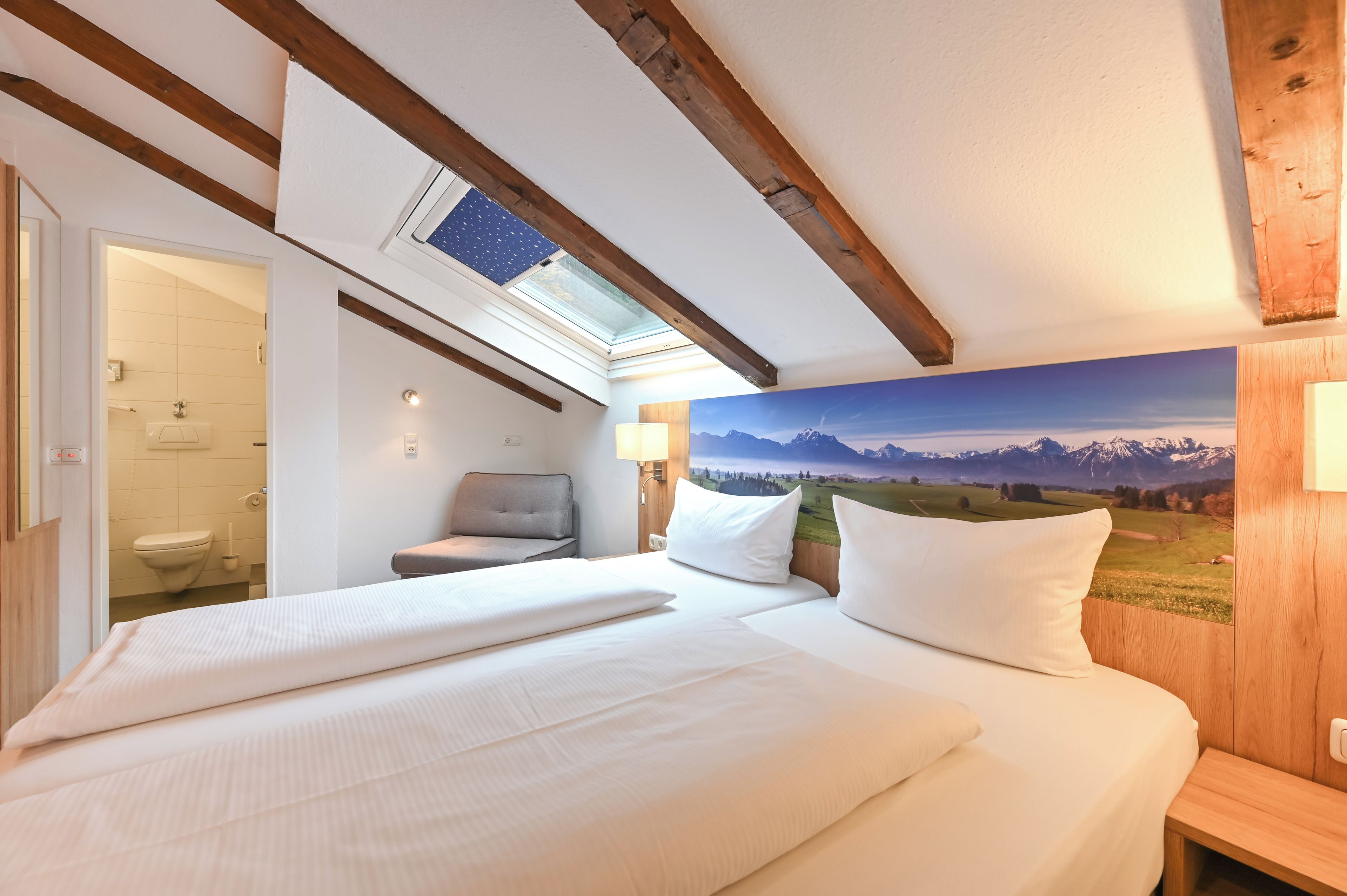 A hotel room with a bed, a chair, a bathroom, and a large window with a mountain view.