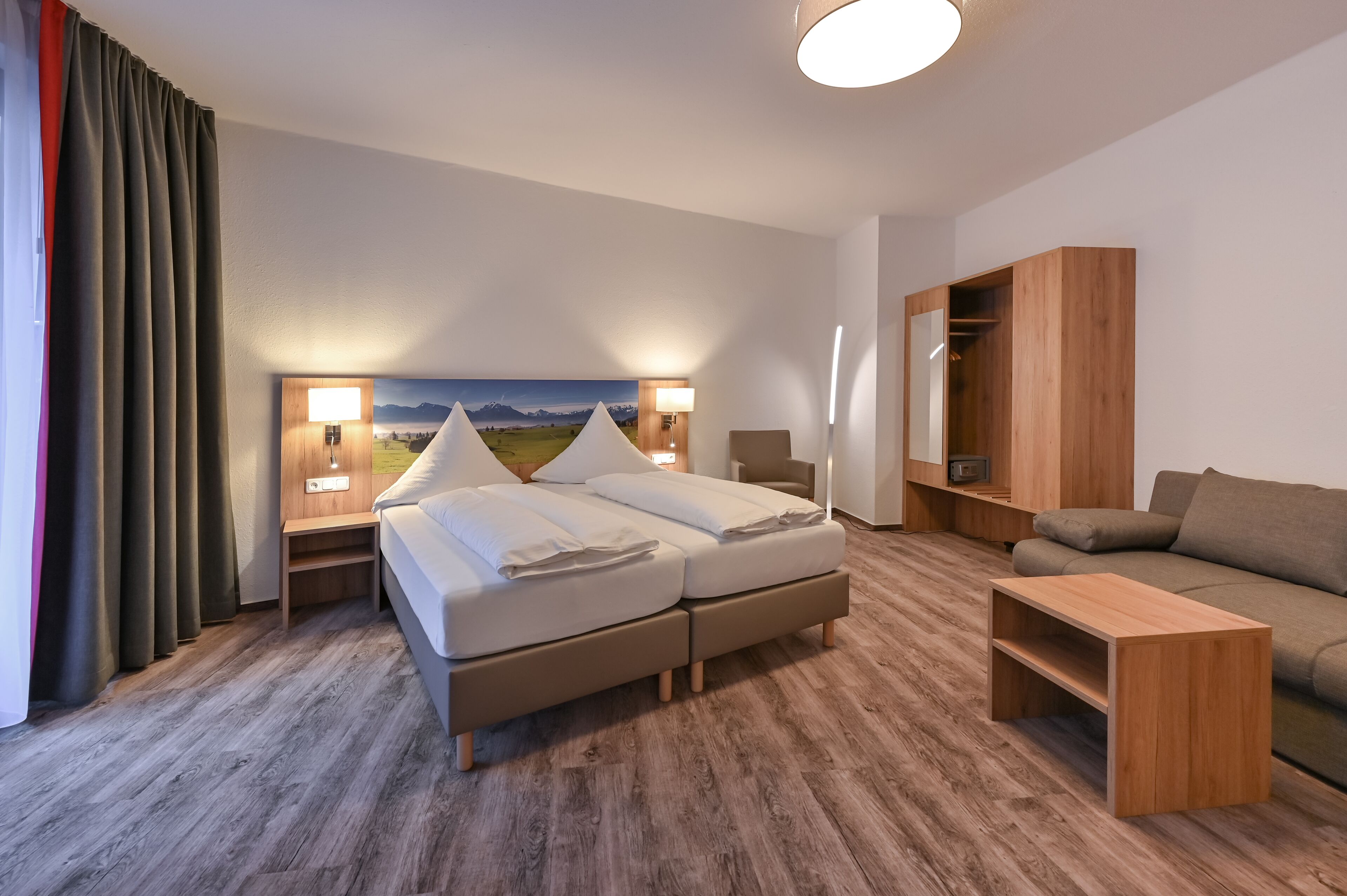A hotel room with a bed, a sofa, a wooden side table, a wardrobe, and a painting on the wall.