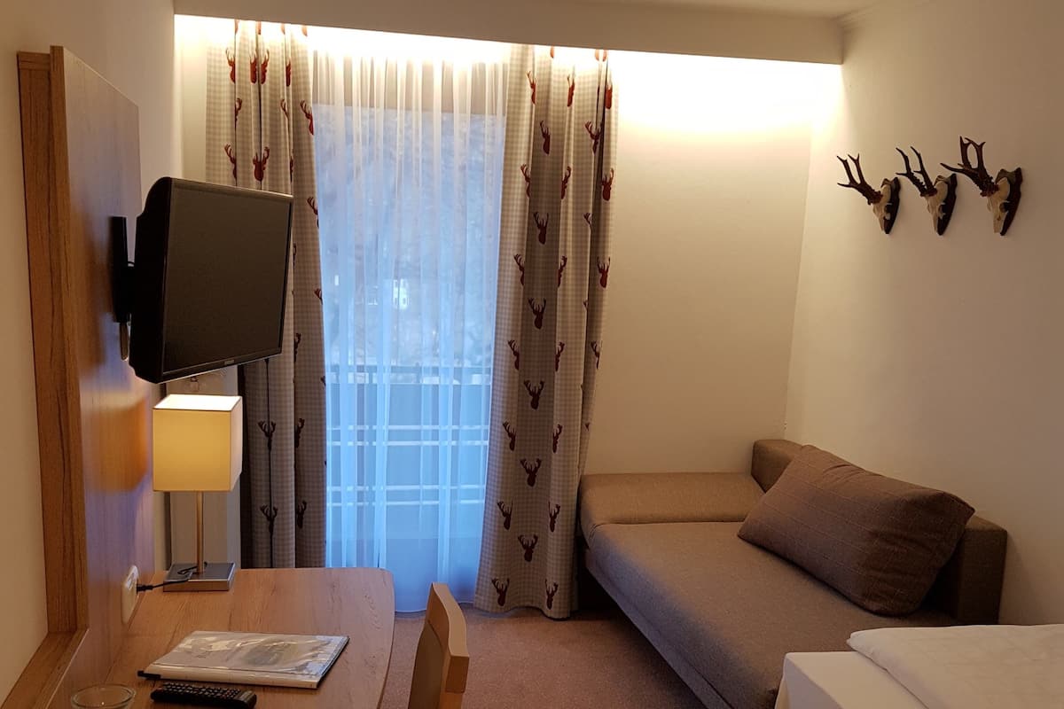A hotel room with a wooden desk, a television mounted on the wall, a sofa, a bed, and a window with curtains.