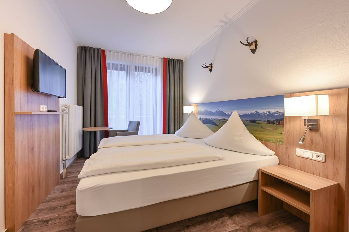 A hotel room with a large bed, a TV mounted on the wall, a desk, and a painting of a mountain landscape.