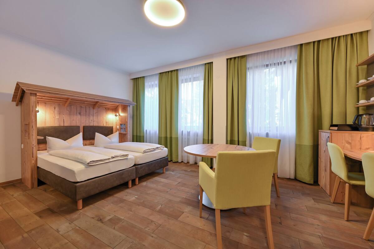 A hotel room with a wooden headboard, two beds, a round dining table with yellow chairs, and a kitchen area with a wooden counter and shelves.