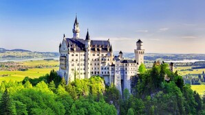 A large, historic castle with multiple towers and a prominent central building, surrounded by lush greenery and a scenic landscape. - Vital Hotel Wiedemann (Fuessen)