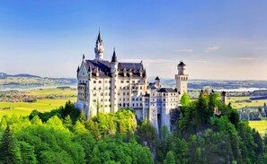 Un castillo grande e histórico con múltiples torres y un edificio central destacado, rodeado de abundante vegetación y un paisaje pintoresco.