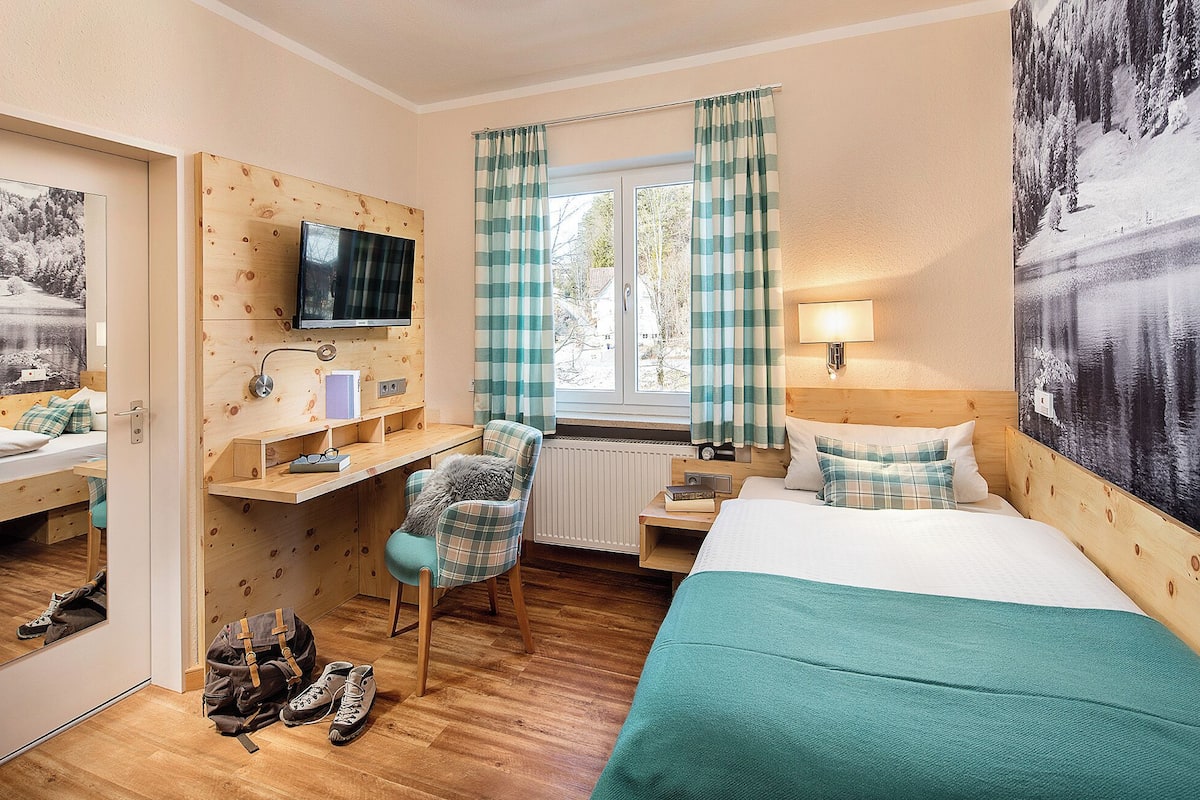 A cozy hotel room with a wooden desk, a bed with a teal bedspread, a chair, and a large window with curtains.