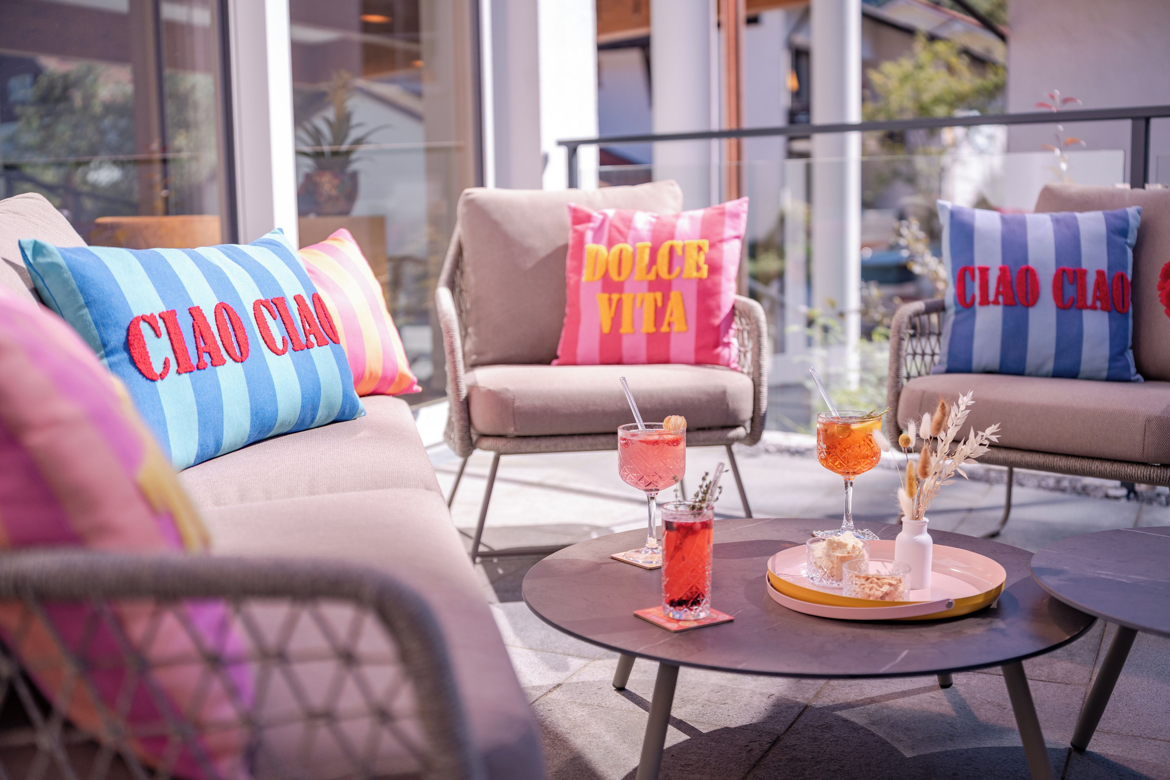 Een zithoek met comfortabele stoelen en kleurrijke kussens, een ronde tafel met drankjes en snacks, en een uitzicht door grote ramen.
