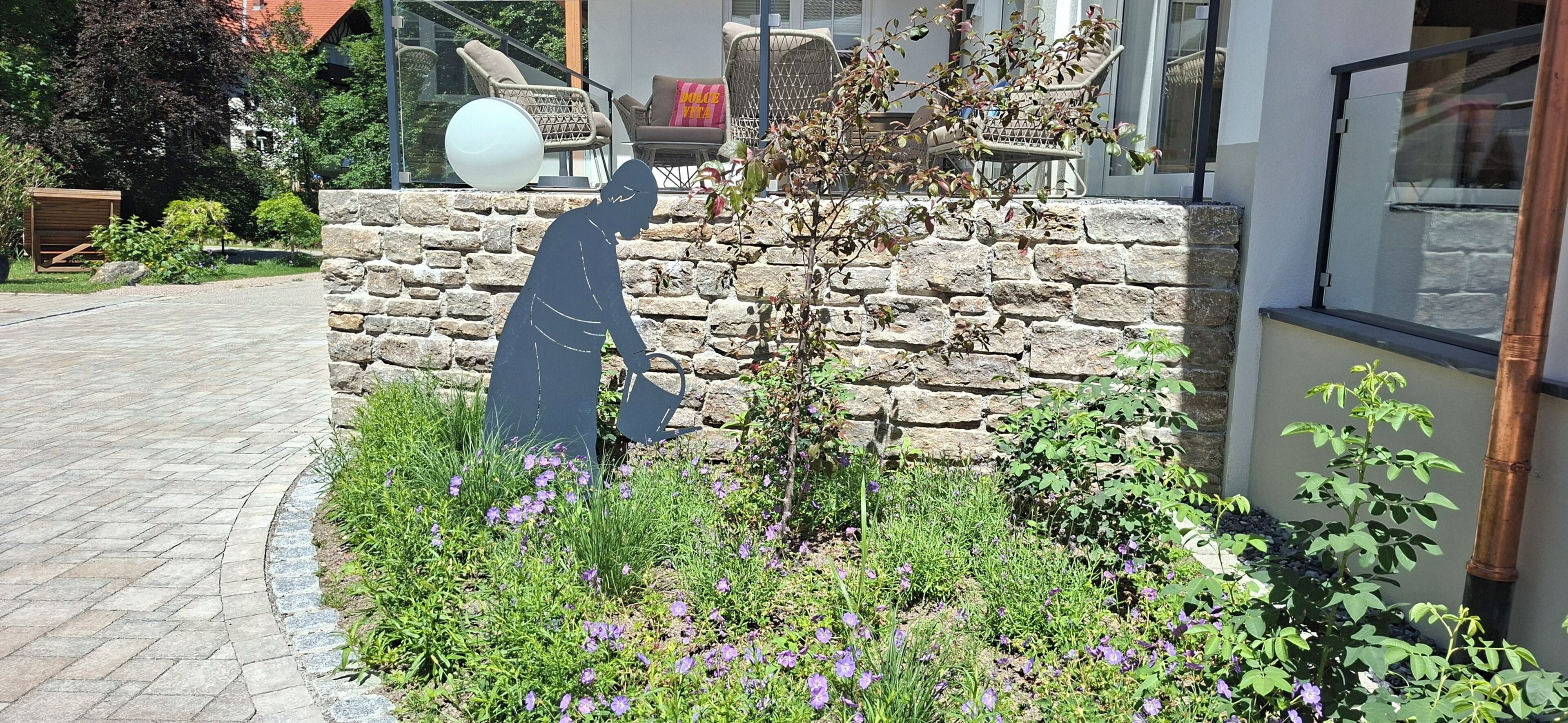 Eine steine Mauer mit einem Garten davor, inklusive violetten Blumen und einem kleinen Baum.