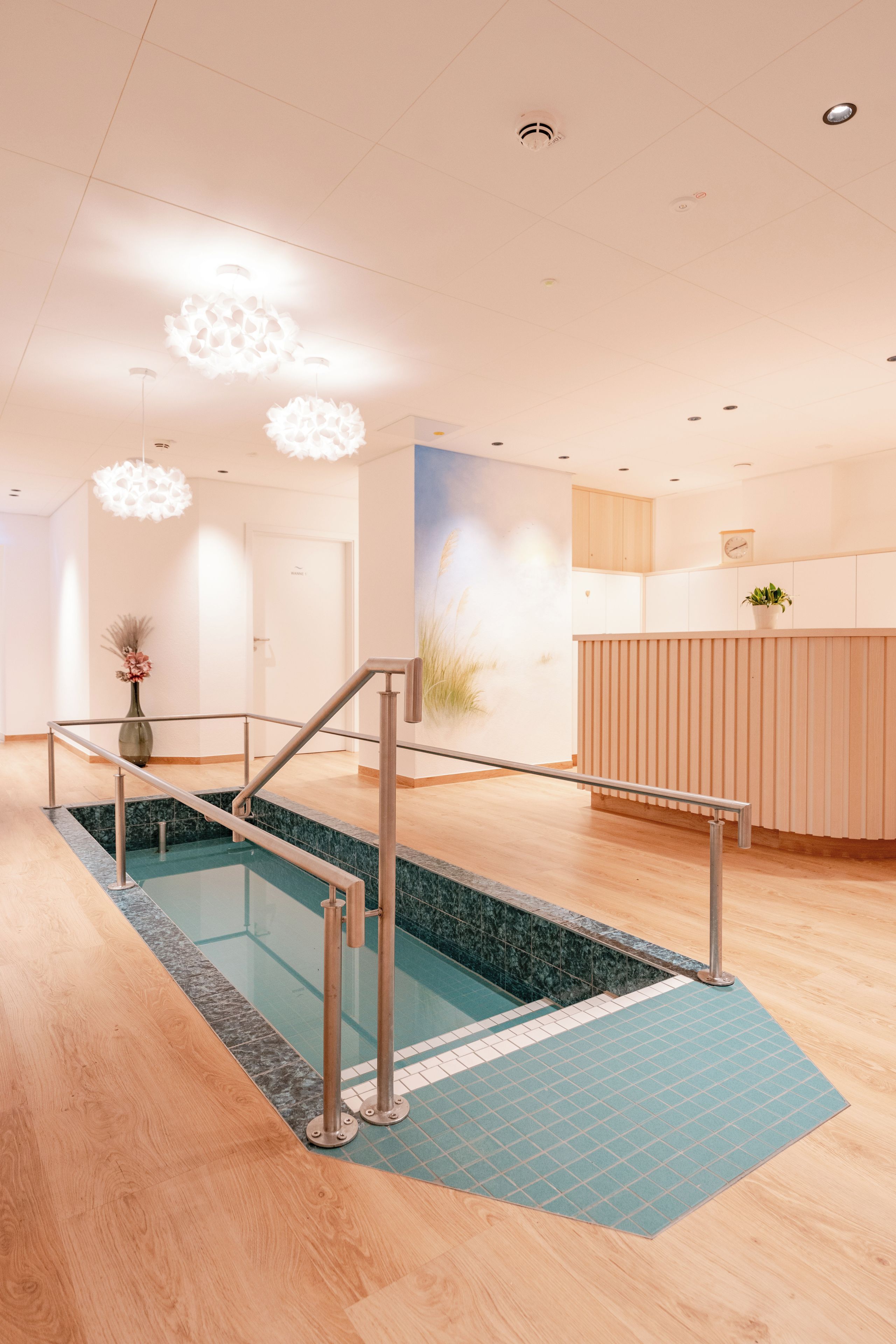 Un moderno vestíbulo de hotel con mostrador de recepción, zona de espera, piscina con barrera de cristal y escalera que conduce al piso superior.