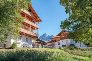 A traditional chalet-style building with a wooden balcony, surrounded by lush greenery and a mountain backdrop. - Vital Hotel Wiedemann (Fuessen)