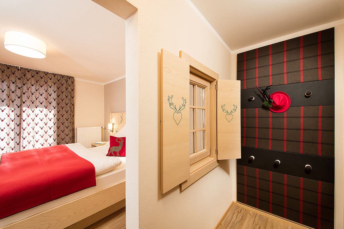A hotel room with a bed, a window with wooden shutters, a red plaid wall panel, and a red hat hanging on the panel.