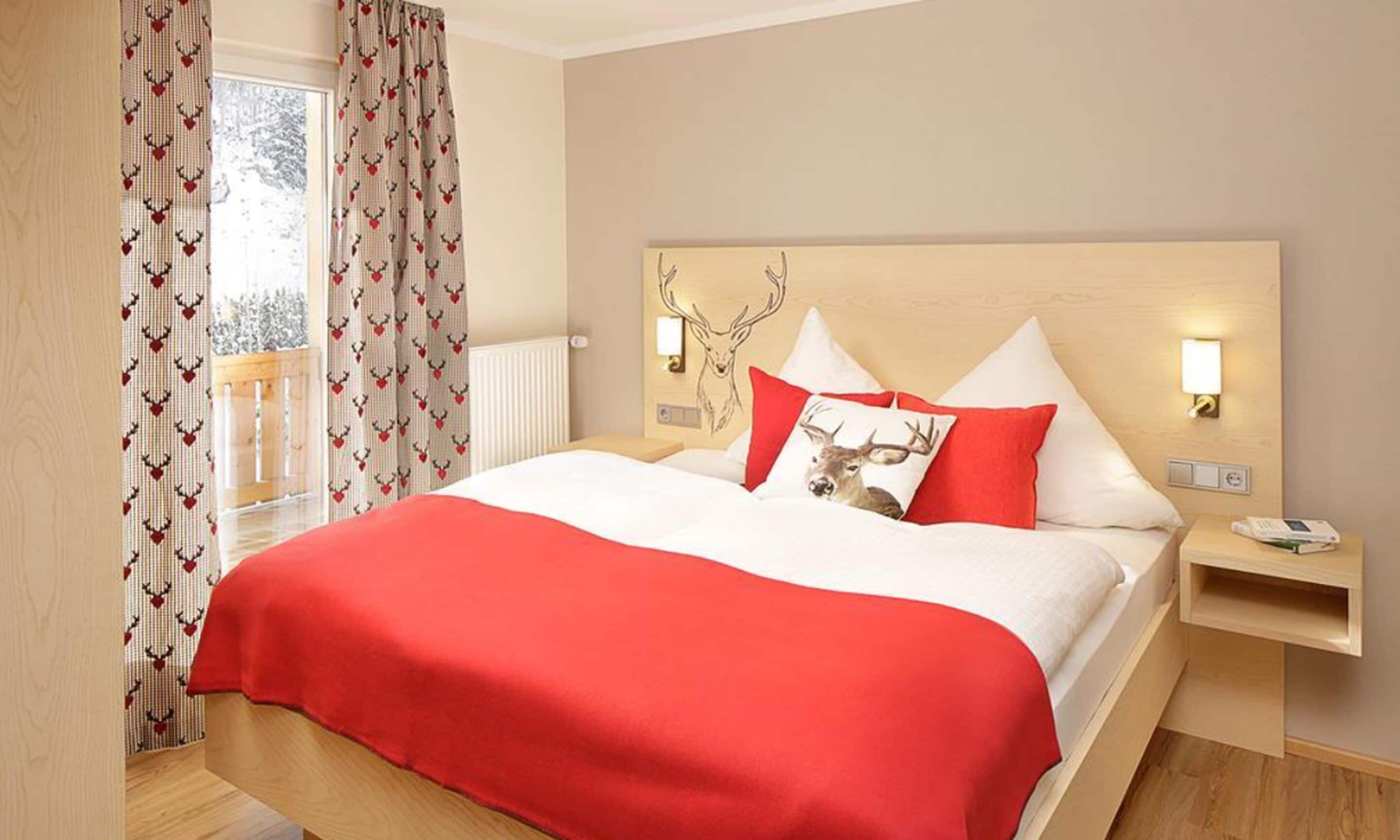 A hotel room with a large bed, a headboard with a deer head design, red and white bedding, a nightstand with a lamp, and a window with curtains.