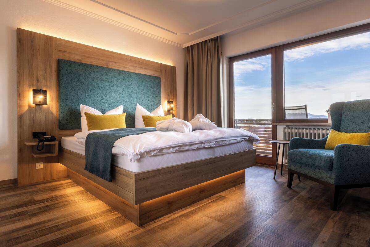Une chambre d’hôtel moderne avec un lit en bois, une petite table, une chaise et une vue sur le ciel grâce à de grandes fenêtres.