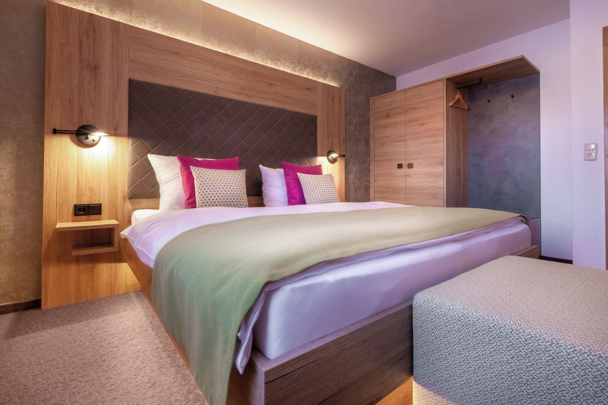 A modern bedroom with a wooden headboard, a bed with a green and white bedspread, two bedside tables with lamps, and a wooden wardrobe.