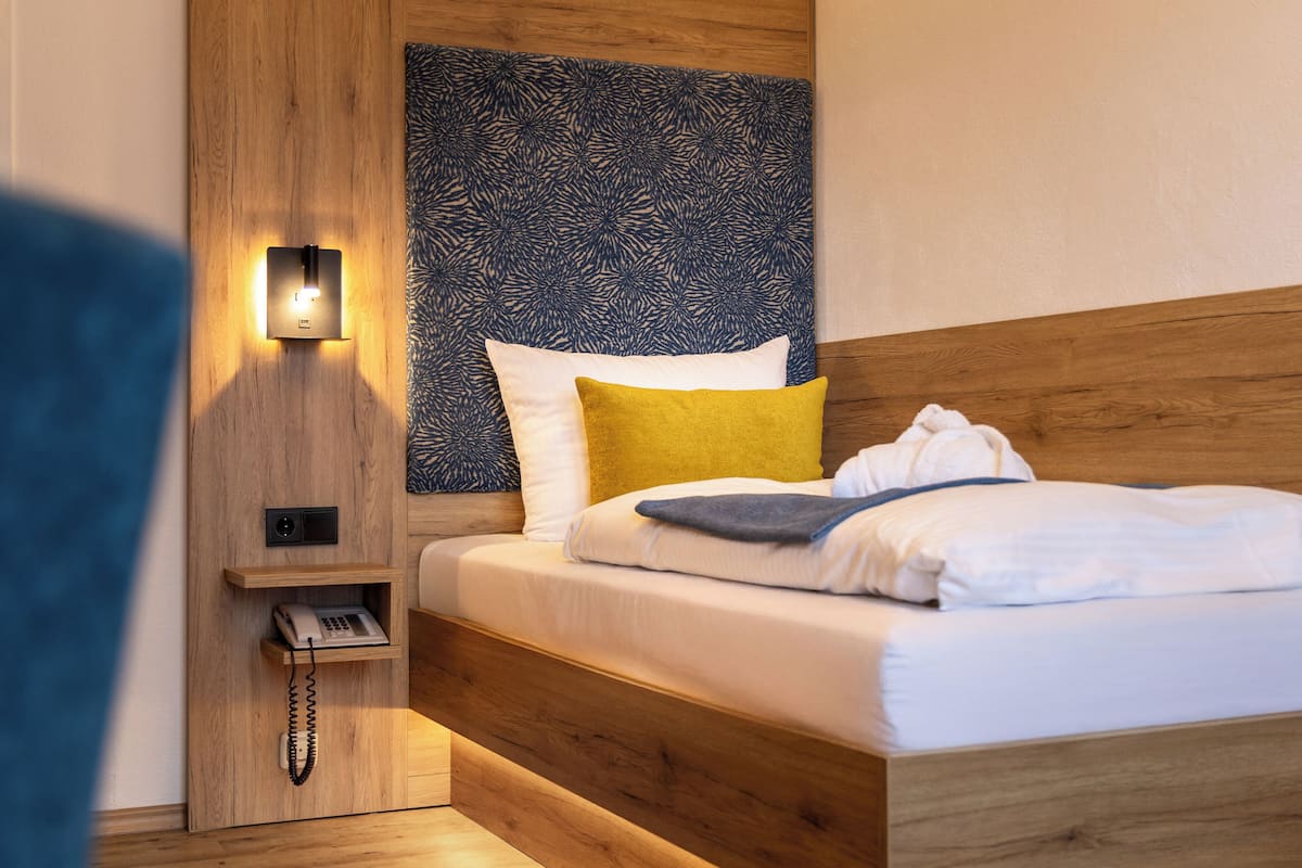Un lit bien fait, avec du linge de lit blanc et un oreiller bleu et jaune, une tête de lit en bois, une table de chevet avec une lampe et un téléphone.