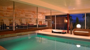 A modern indoor swimming pool with clear glass walls, a wooden deck, and a seating area with a large round table and chairs. - Hotel Bergruh (Fuessen)