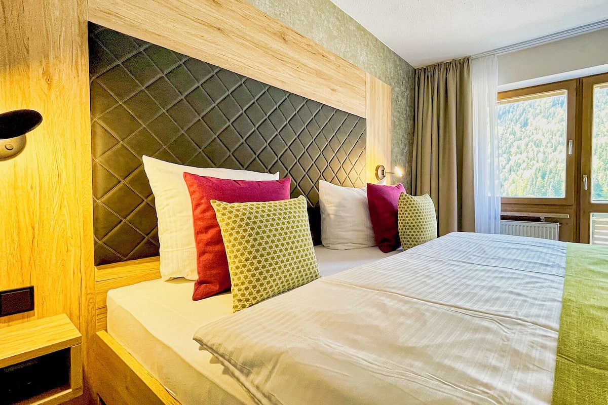 Une chambre d’hôtel avec un grand lit, plusieurs oreillers, une tête de lit à motifs losangés, une table de chevet et une fenêtre donnant sur des arbres.