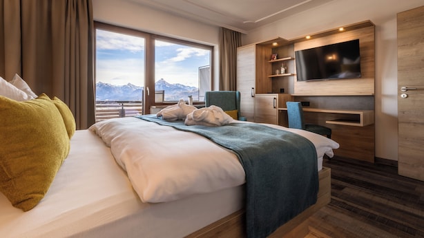 A modern hotel room with a large bed, a TV mounted on the wall, and a view of mountains through the window. - Hotel Bergruh (Fuessen)