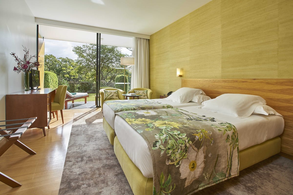 A modern hotel room with a large bed, a desk, and a chair. There is a view of the outdoors through large windows.