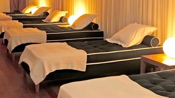 Varias camillas de masaje alineadas, cubiertas con sábanas blancas y almohadas, en una habitación con suelo de madera y cortinas.