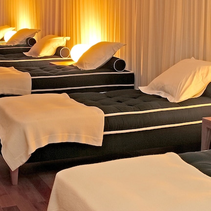 A series of massage beds arranged in a row with white linens and pillows, in a room with wooden flooring and curtains.