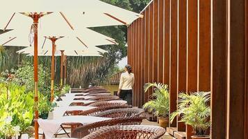 A row of wicker tables and chairs with white tablecloths, shaded by large umbrellas, along a wooden wall with vertical slats.