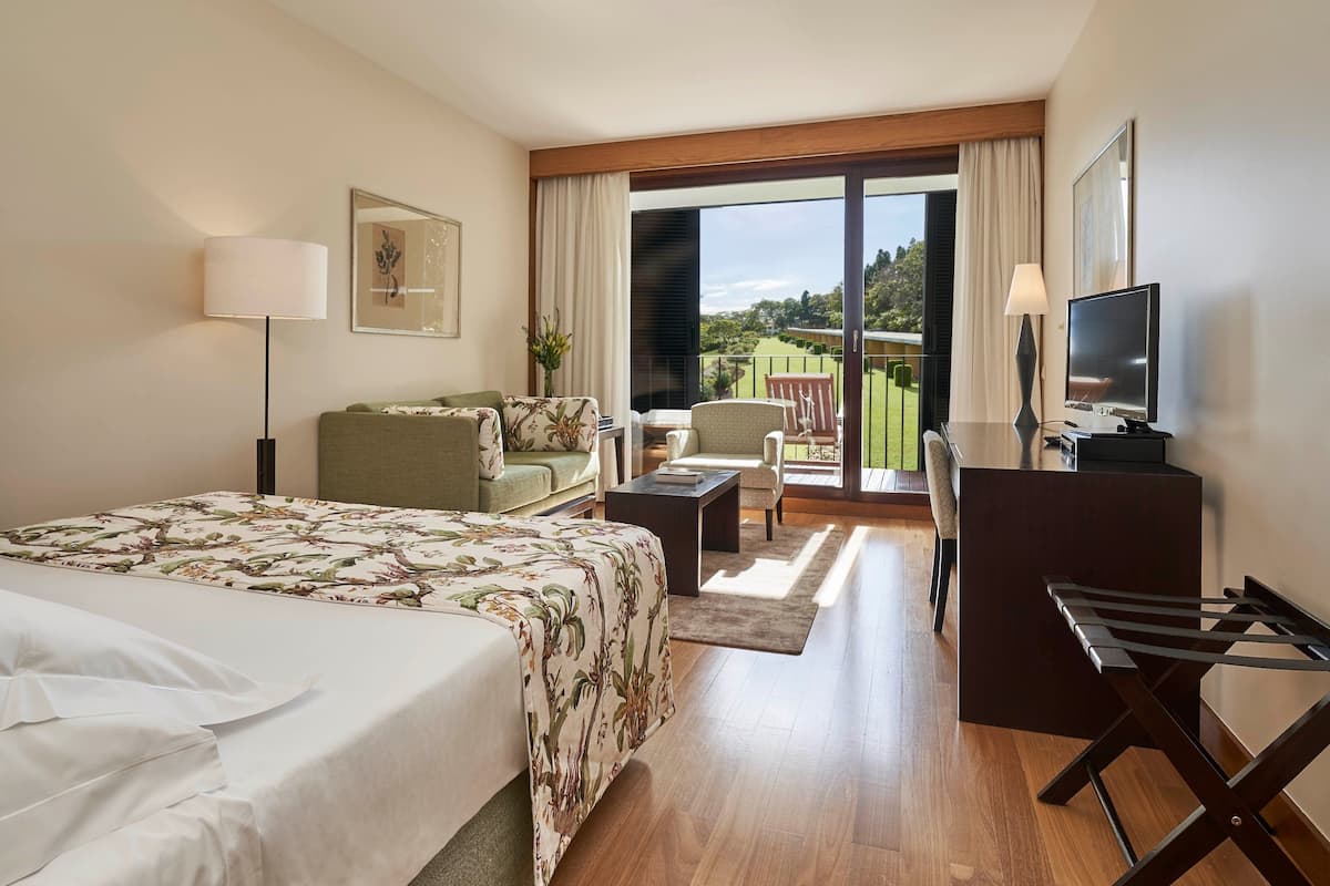 A hotel room with a bed, a desk, a chair, a sofa, a TV, and a balcony with a view of the outdoors.
