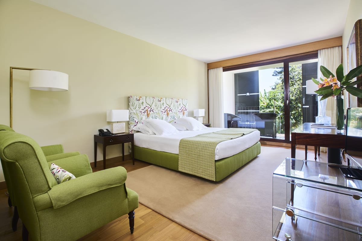 A hotel room with a bed, a green armchair, a glass table, a nightstand, and a view of the outdoors.