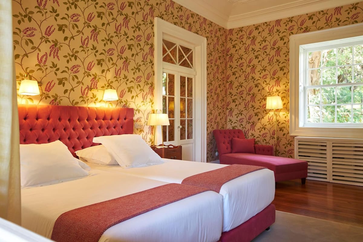 A bedroom with a large bed, two bedside tables, a red upholstered chair, and a window with a view of greenery.