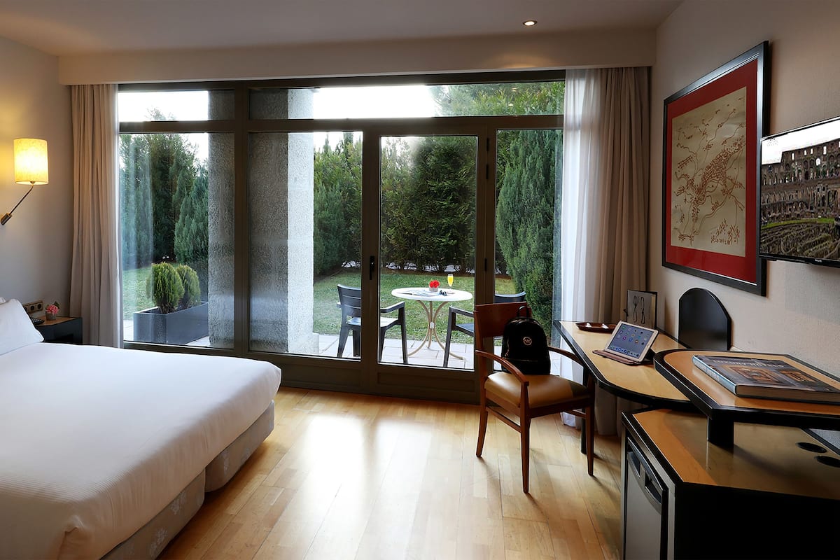 Habitació d'hotel amb un llit, una taula de treball amb un ordinador portàtil, una cadira i vista a un jardí a través d'una finestra gran.